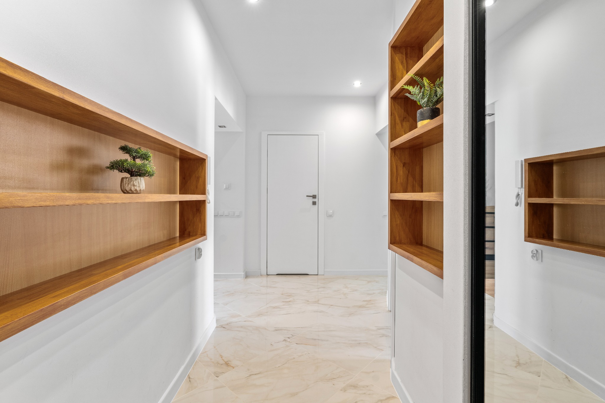 Nowoczesny korytarz z drewnianymi półkami i marmurową podłogą. W lustrze odbija się fragment korytarza z identycznymi półkami. Minimalistyczny design.