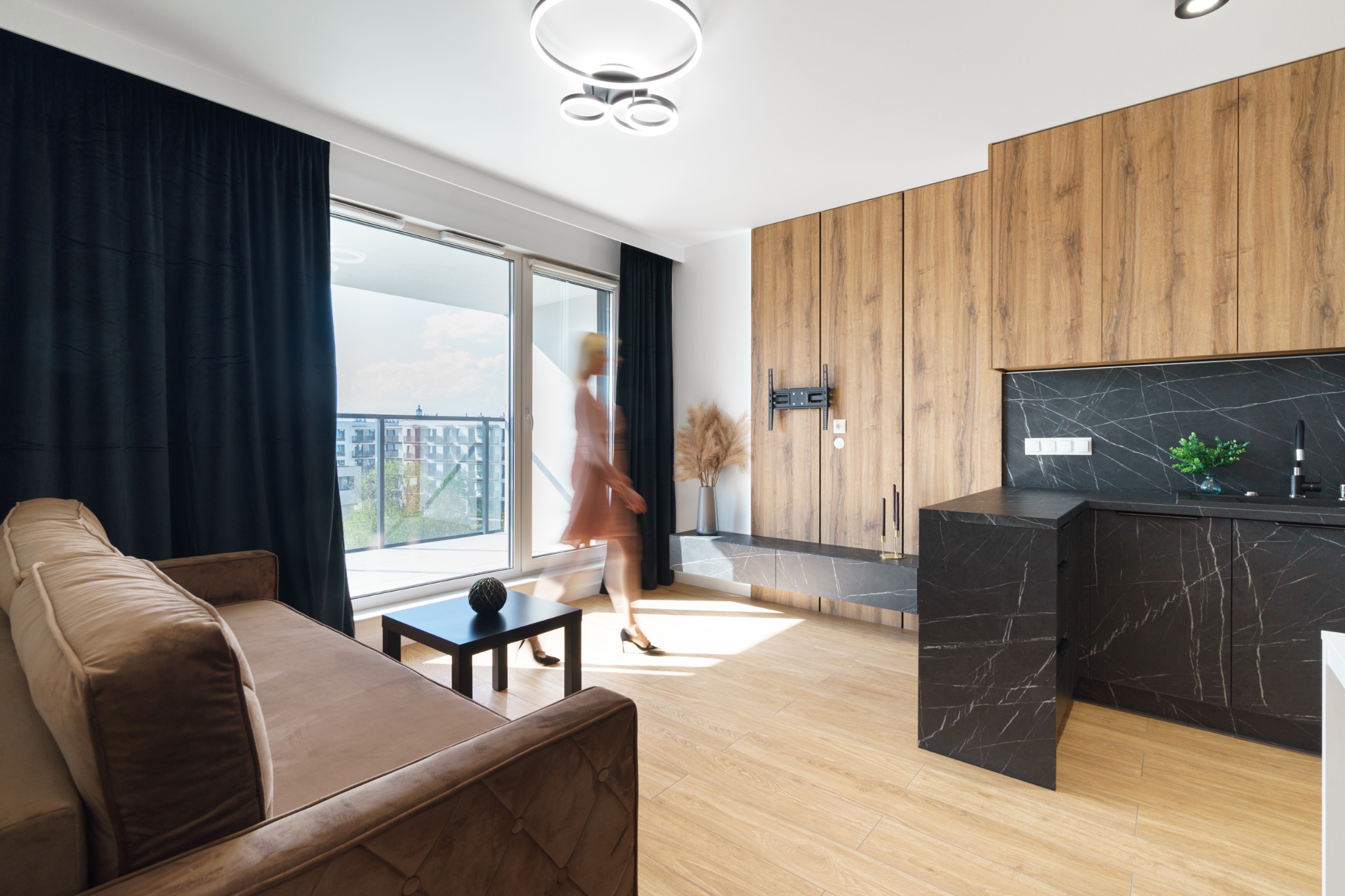 Nowoczesne wnętrze: sofa, stół, zabudowa drewniana z TV, marmurowy blat kuchenny. Rozmyta sylwetka kobiety przechodzącej obok balkonu. Stylowe oświetlenie sufitowe.