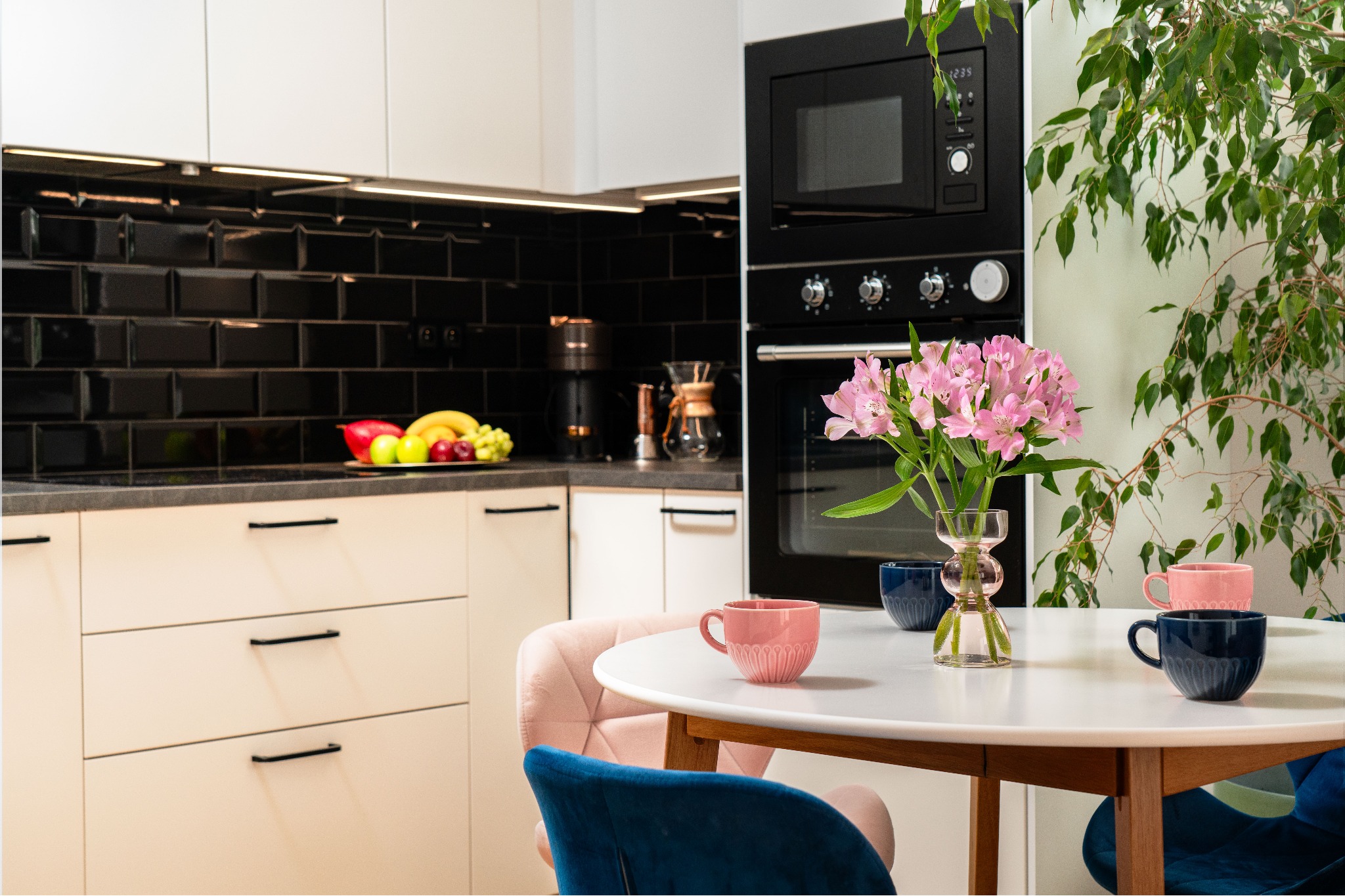 Nowoczesna kuchnia z czarnymi płytkami, białymi szafkami i okrągłym stołem z kwiatami w wazonie. Aranżacja wnętrza z dbałością o detale.