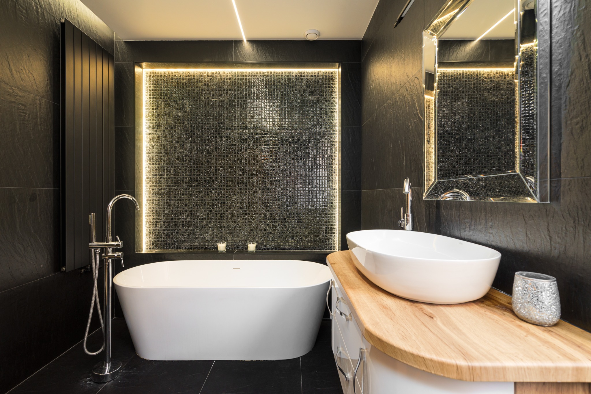 Ekskluzywna łazienka z czarnymi płytkami, wolnostojącą białą wanną, umywalką na drewnianym blacie i podświetlaną mozaiką. Nowoczesny design i eleganckie wykończenie.