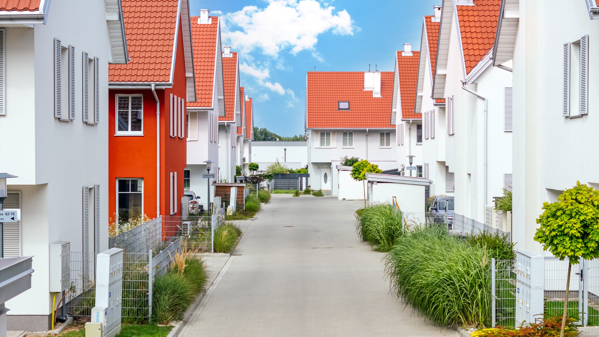 Ulica z rzędem nowoczesnych domów w jasnych kolorach z czerwoną dachówką, zielenią i błękitnym niebem w tle. Jeden dom wyróżnia się pomarańczową elewacją.