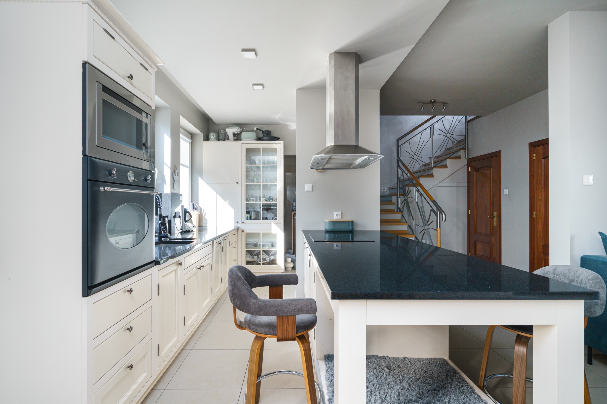 Jasna kuchnia z wyspą i hokerami, widok na schody. Białe szafki, czarny blat, nowoczesny okap. W tle schody z drewnianymi stopniami i metalową balustradą.