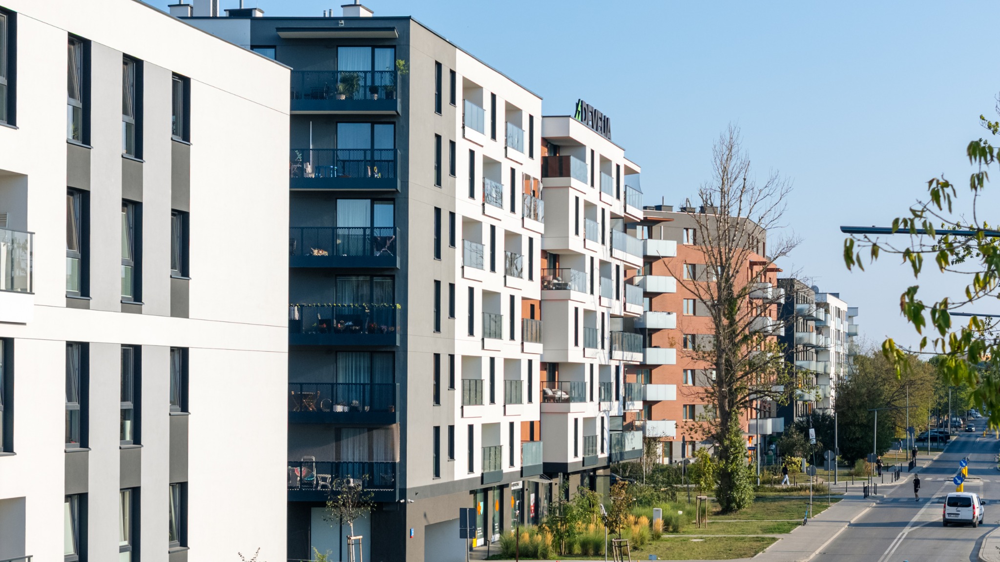 Nowoczesny kompleks apartamentów z balkonami i przeszkleniami, widok z perspektywy ulicy, zieleń w tle, błękitne niebo. Budynki o różnej kolorystyce elewacji.