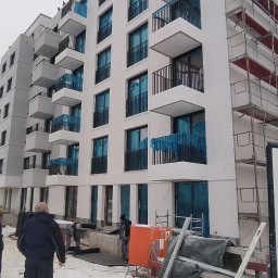 Flota Investment - Nowoczesny, biały budynek wielorodzinny z balkonami i rusztowaniami. Na pierwszym planie pracownicy wykonujący prace wykończeniowe na zewnątrz budynku.