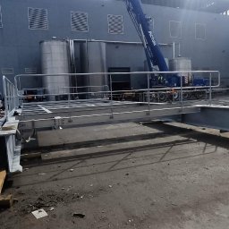 Flota Investment - Montaż konstrukcji stalowej z użyciem dźwigu na tle przemysłowego budynku. Widoczni pracownicy w kaskach i kamizelkach odblaskowych. Elementy konstrukcyjne na ziemi.