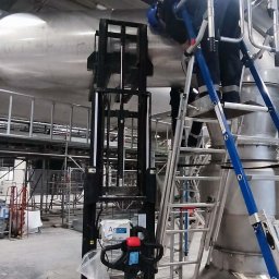 Flota Investment - Pracownik na drabinie przy metalowej konstrukcji w hali przemysłowej, obok podnośnik. Widoczne rury, rusztowania i elementy instalacji. Prace montażowe w toku.