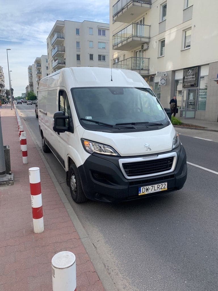 Biały bus marki Peugeot zaparkowany na ulicy w mieście, z widocznymi budynkami mieszkalnymi w tle i słupkami drogowymi w kolorze biało-czerwonym.
