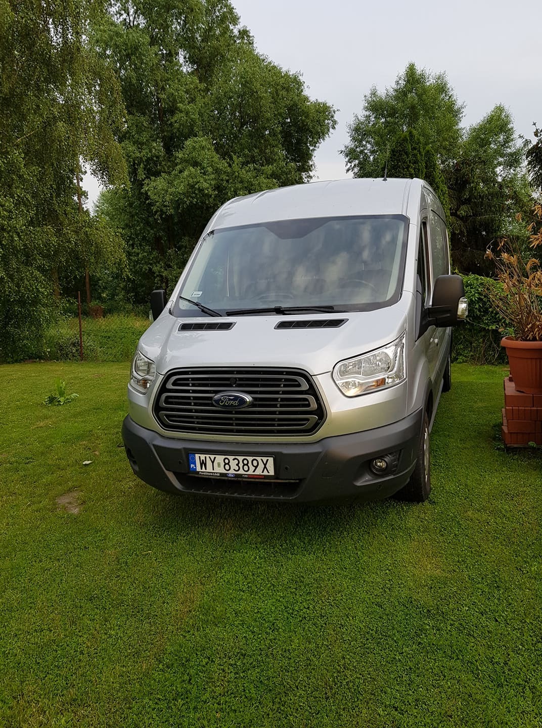 Srebrny Ford Transit na trawniku, z widoczną tablicą rejestracyjną WY 8389X, drzewa w tle.