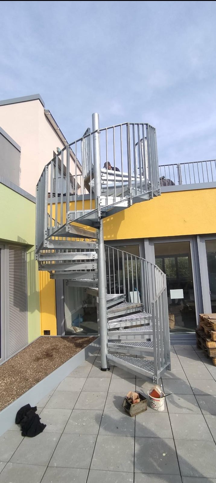 Zewnętrzne, metalowe schody spiralne prowadzące na taras budynku z żółtą elewacją. Widoczne narzędzia i materiały budowlane. Jasne, naturalne oświetlenie.