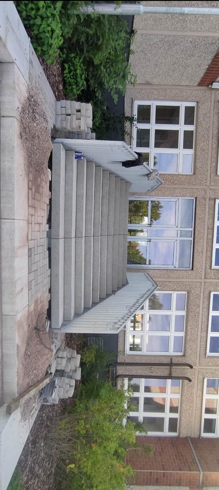 Nowe, betonowe schody zewnętrzne z metalową barierką, prowadzące do budynku z oknami. Częściowo ułożona kostka brukowa i stos kostek obok schodów.