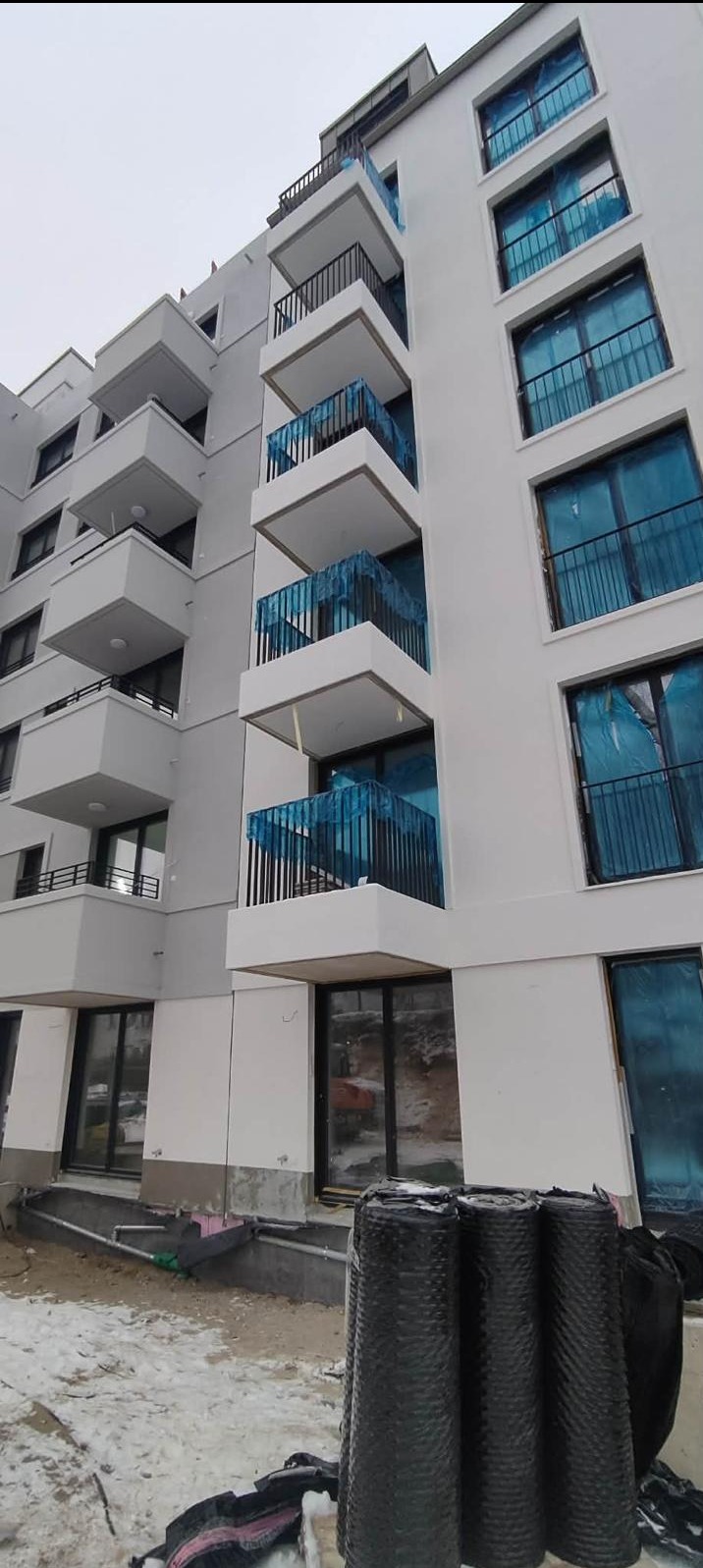 Nowoczesny budynek wielorodzinny z balkonami zabezpieczonymi folią w Warszawie. Widoczne rolki materiału izolacyjnego, sugerujące prace wykończeniowe.