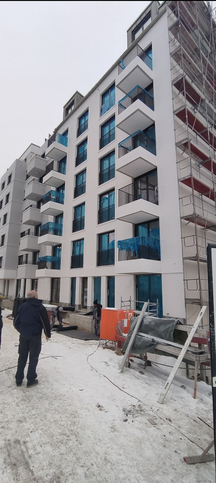 Nowoczesny, biały budynek wielorodzinny z balkonami i rusztowaniami. Na pierwszym planie pracownicy wykonujący prace wykończeniowe na zewnątrz budynku.