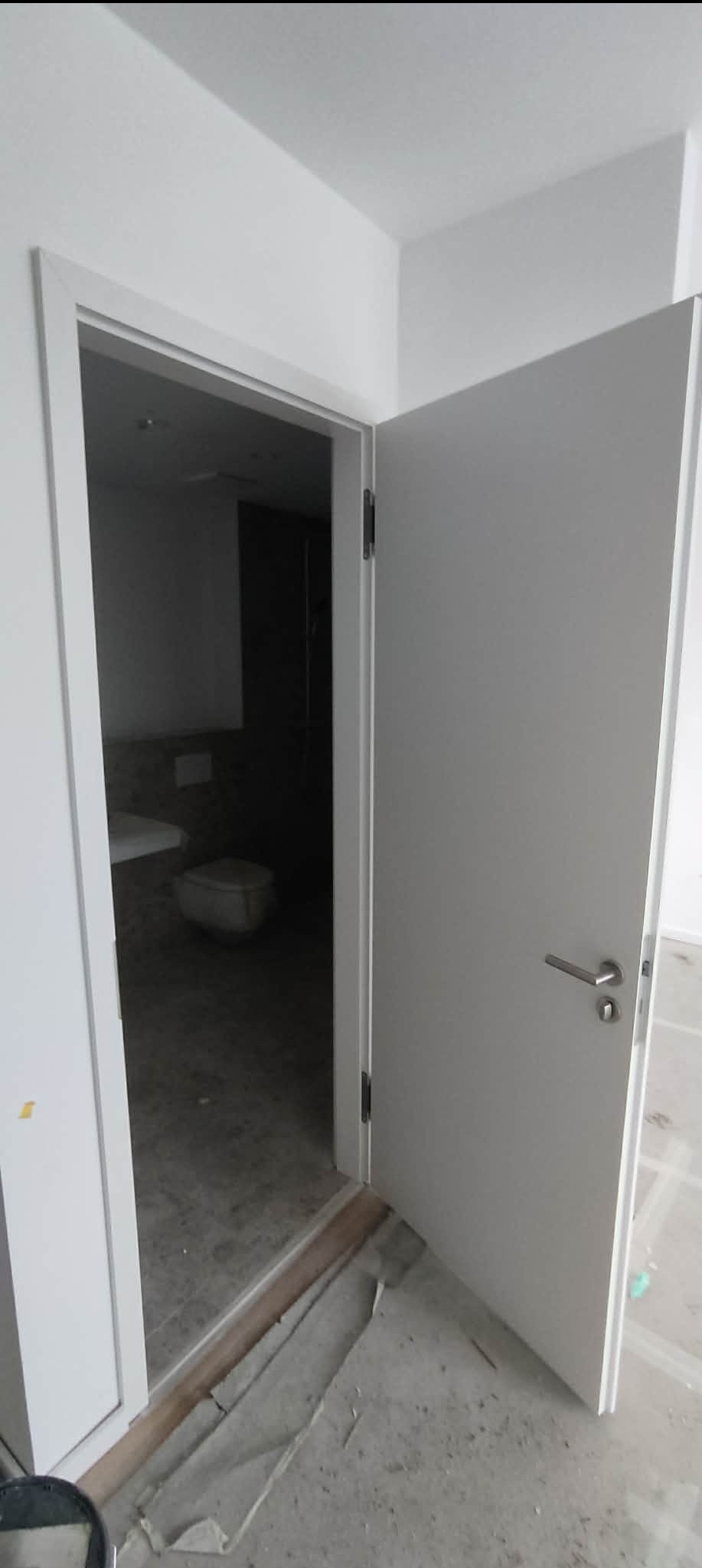 Otwarta, biała, nowoczesna wewnętrzna drzwi z widokiem na łazienkę w trakcie remontu. Widoczne zabezpieczenia podłogi i surowy stan wykończenia.