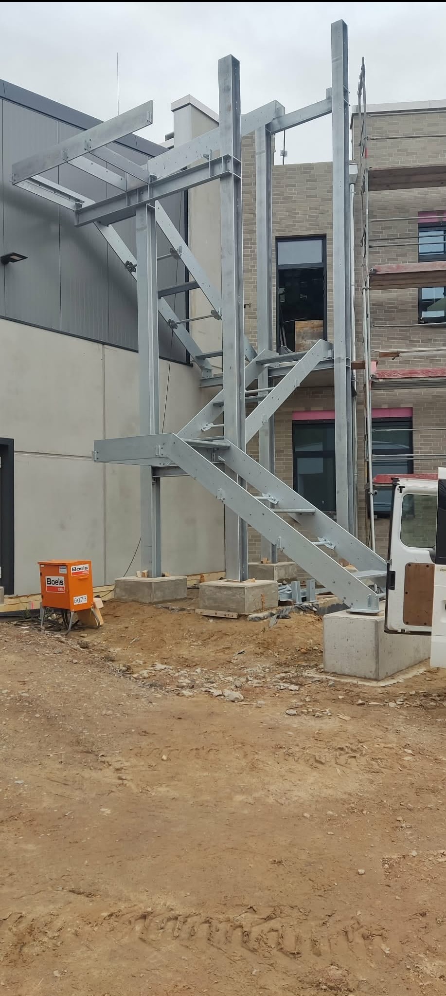 Stalowa konstrukcja schodów zewnętrznych na betonowych fundamentach, częściowo zamontowana przy budynku z cegły i szarym panelowym wykończeniem.