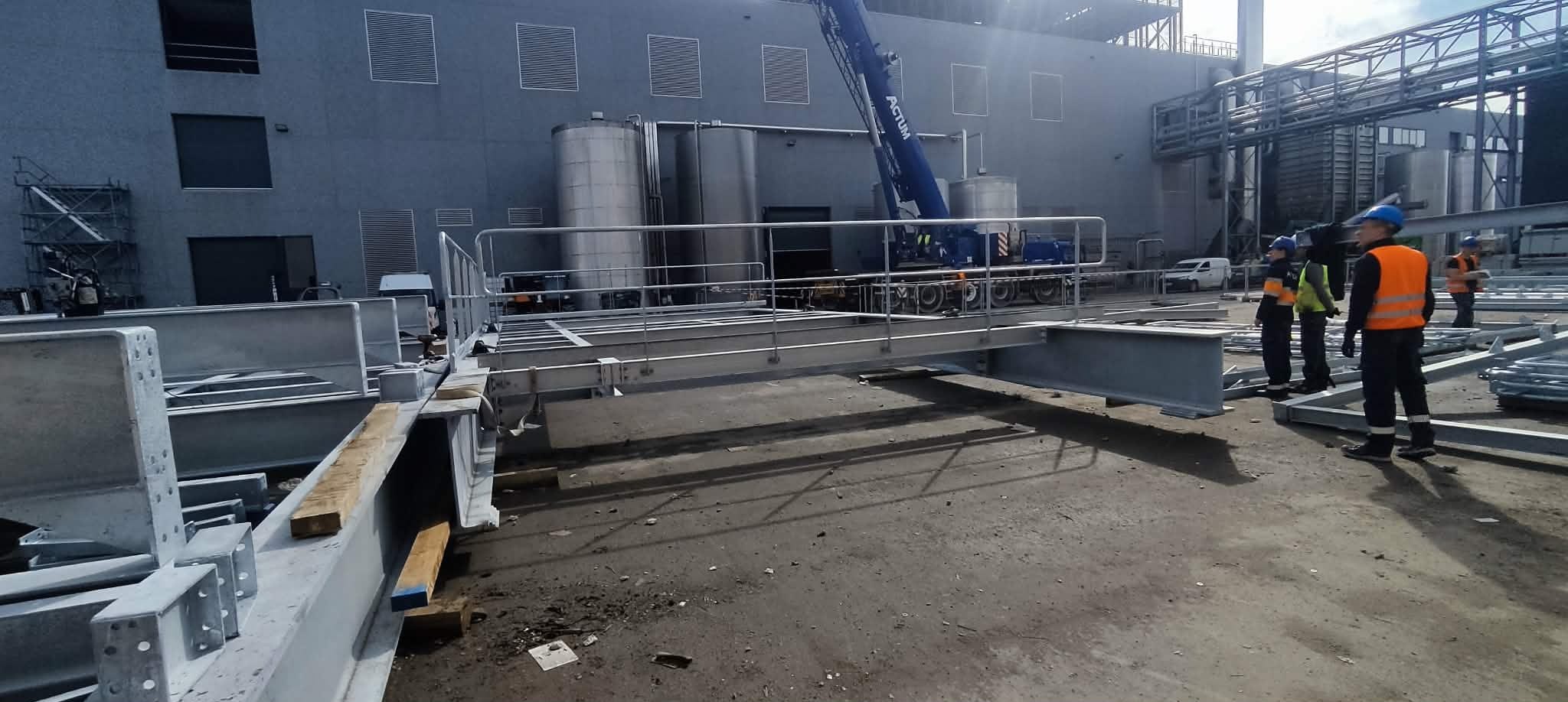 Montaż konstrukcji stalowej z użyciem dźwigu na tle przemysłowego budynku. Widoczni pracownicy w kaskach i kamizelkach odblaskowych. Elementy konstrukcyjne na ziemi.