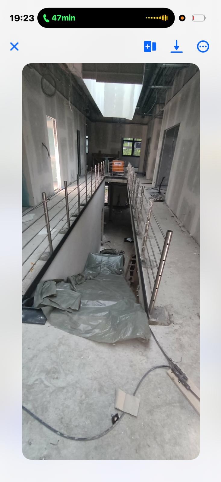 Wnętrze w trakcie remontu: surowe ściany, metalowe barierki schodowe, zabezpieczona folią podłoga. Widoczne okno i elementy instalacji pod sufitem. Surowy stan wykończenia.