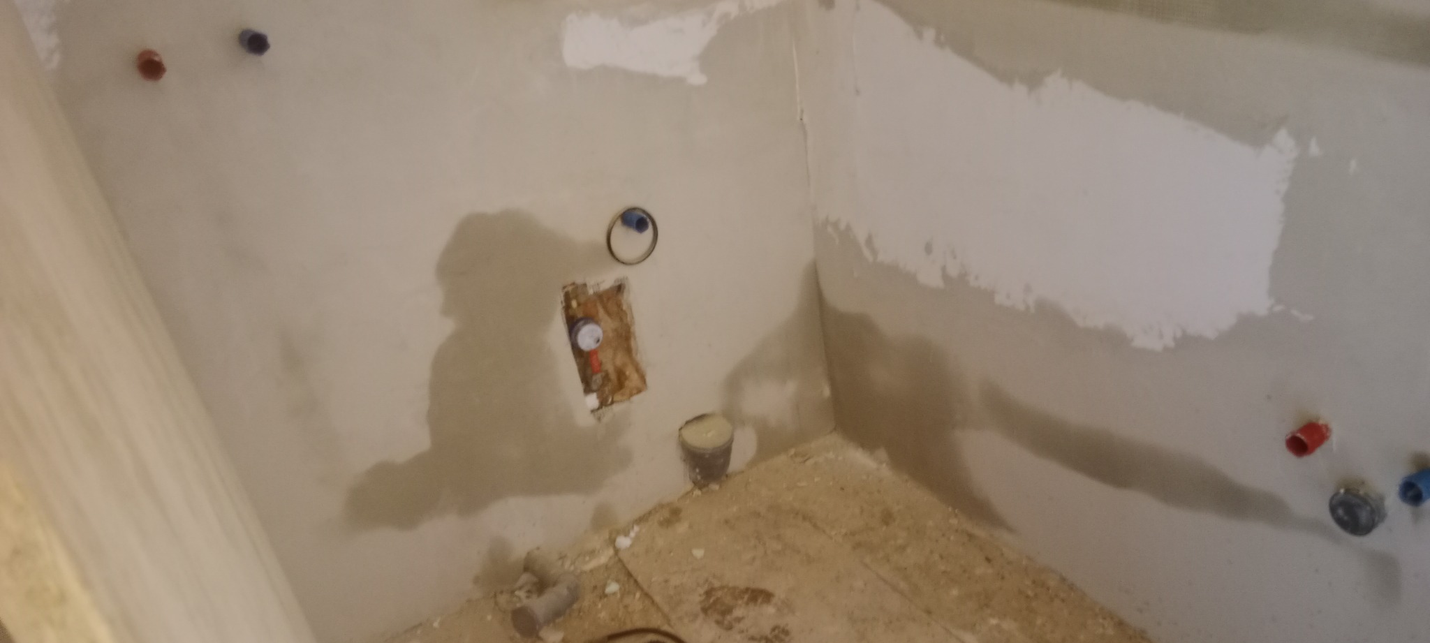 Surowy stan łazienki w trakcie remontu: ściany po gruntowaniu, widoczne rury instalacji wodnej z czerwonymi i niebieskimi zaślepkami, przygotowane miejsce na baterię, podłoga z resztkami materiałów...