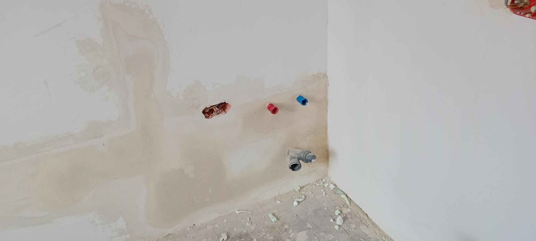 Ściana w trakcie prac wykończeniowych z widocznymi otworami na instalacje elektryczne i wodne, częściowo pokryta gładzią, z fragmentami pianki montażowej na podłodze.