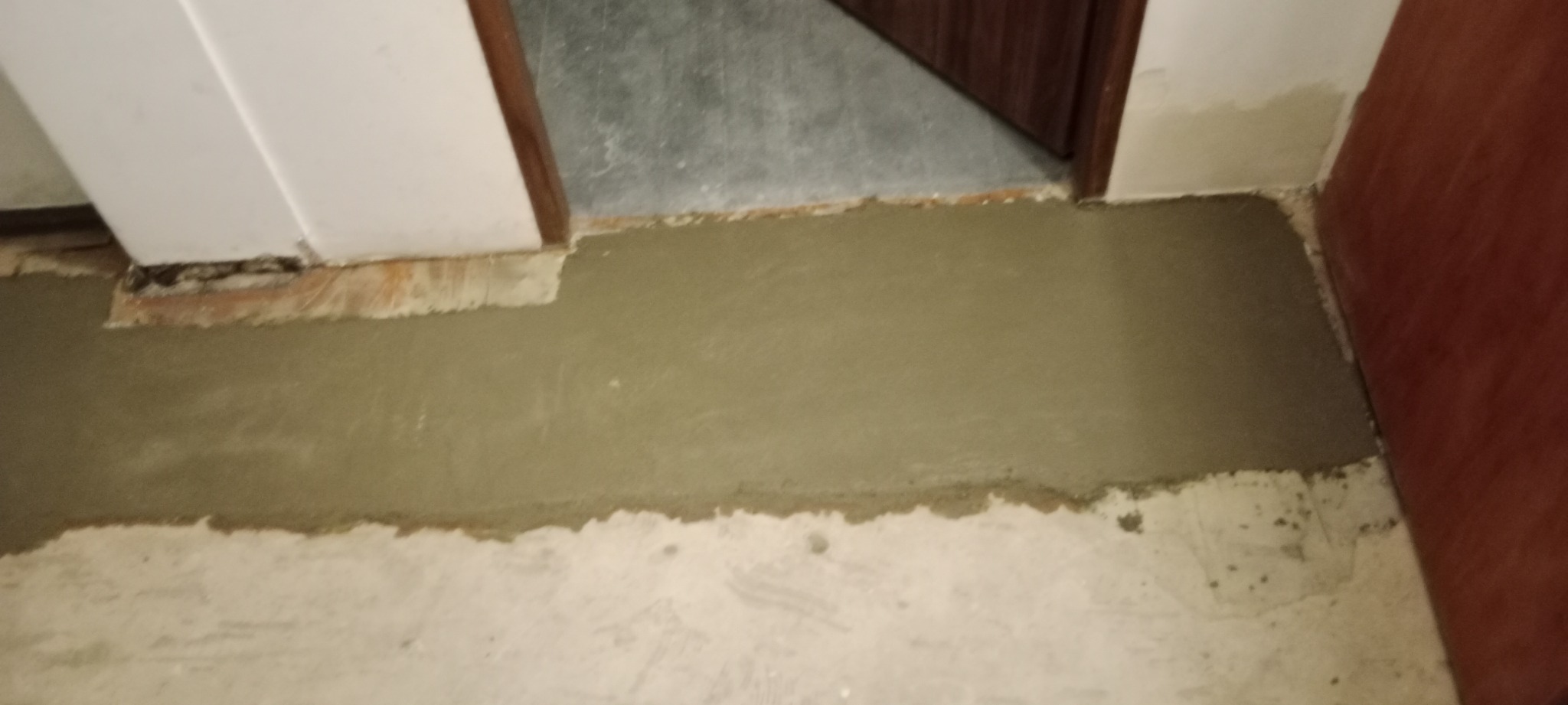 Wyrównywanie podłoża w pomieszczeniu za pomocą wylewki samopoziomującej, widoczne fragmenty ściany i drzwi.