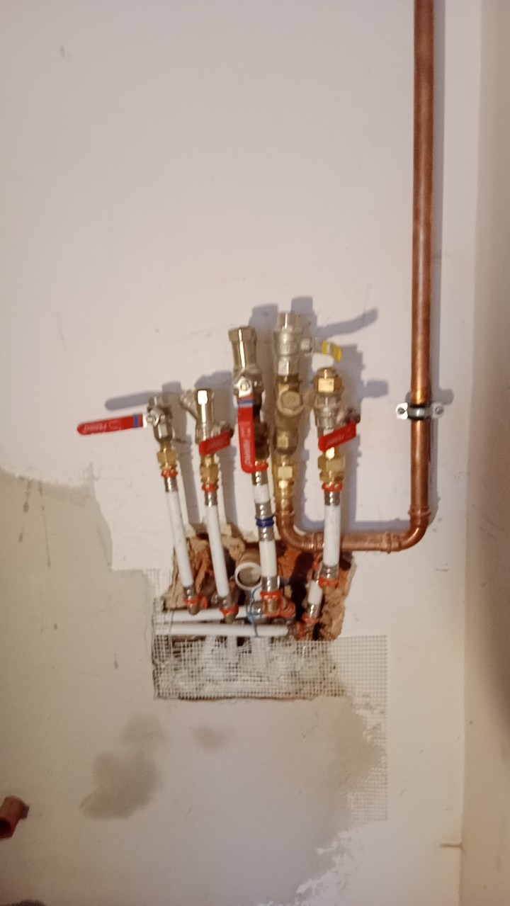 Rozdzielacz wodny z zaworami odcinającymi i rurami miedzianymi, zamontowany na ścianie z widocznym fragmentem siatki do tynkowania i śladami po pracach instalacyjnych.