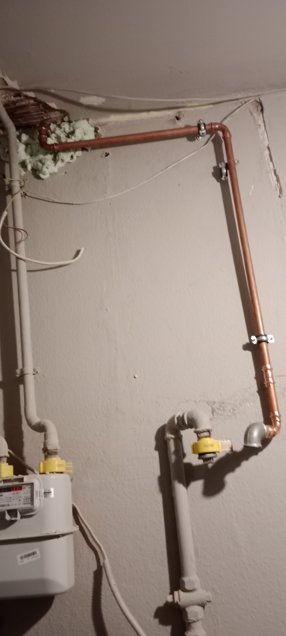 Miedziana instalacja gazowa z licznikiem, częściowo zamaskowana pianką montażową przy suficie, przymocowana do ściany za pomocą metalowych uchwytów.