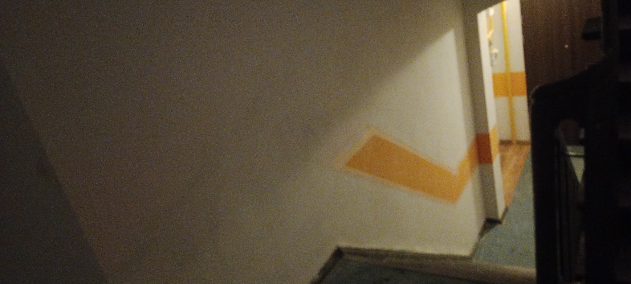 Fragment ściany po malowaniu z widocznymi śladami taśmy malarskiej, w tle fragment korytarza z pomalowanymi paskami.