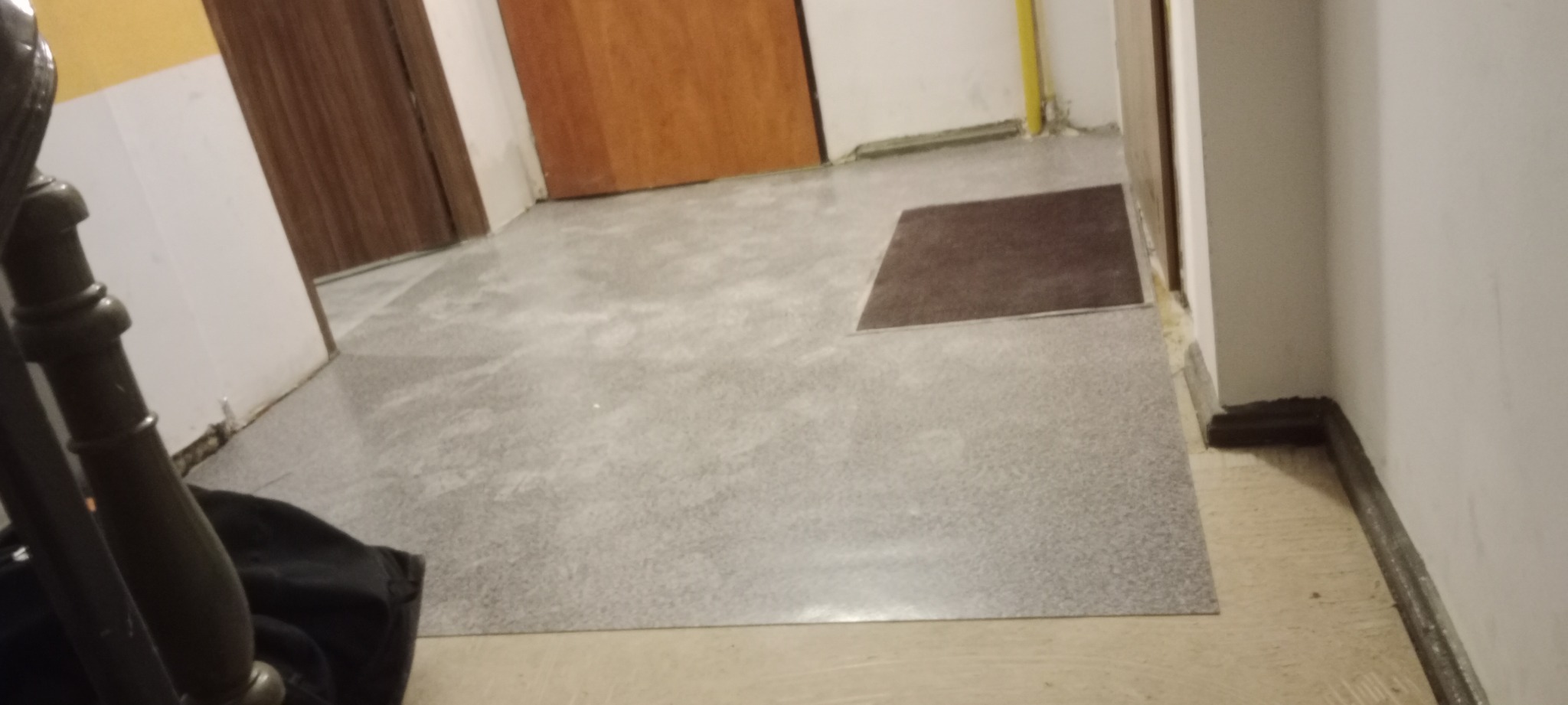 Szary, wypolerowany beton na podłodze w korytarzu, z brązowym dywanikiem przy wejściu do pokoju. Widoczne fragmenty drewnianych drzwi i żółtej rury przy ścianie.