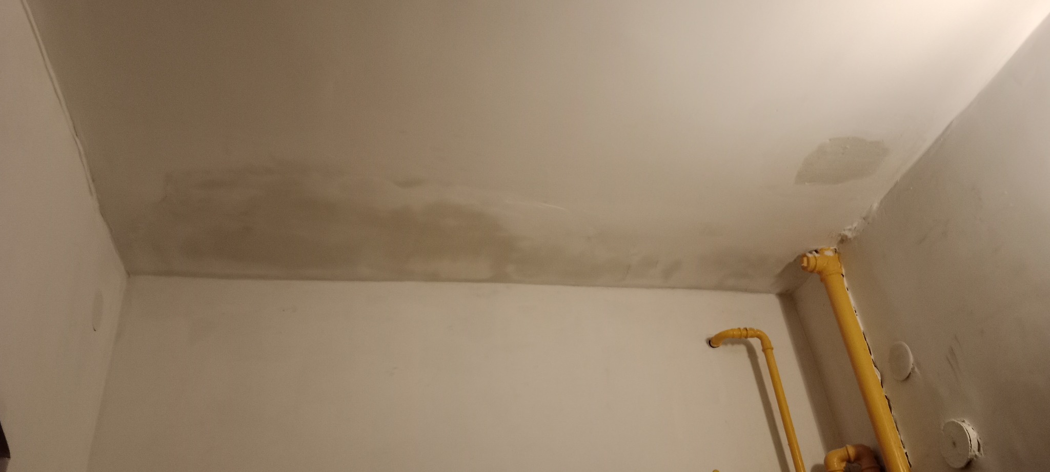 Niedokończone malowanie sufitu z widocznymi zaciekami i żółtymi rurami instalacji gazowej przy ścianie.
