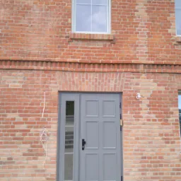 Szare drzwi wejściowe z bocznym przeszkleniem wkomponowane w czerwoną ceglaną elewację budynku z widocznymi oknami i instalacją elektryczną na ścianie.