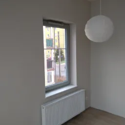 Wnętrze pokoju z oknem z widokiem na blok, biały grzejnik pod oknem, drewniana podłoga i papierowa lampa wisząca.