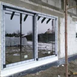 Nowe okno przesuwne z PCV z szybami refleksyjnymi, zamontowane w surowym budynku w trakcie budowy, widoczne białe profile okienne z naklejkami producenta.