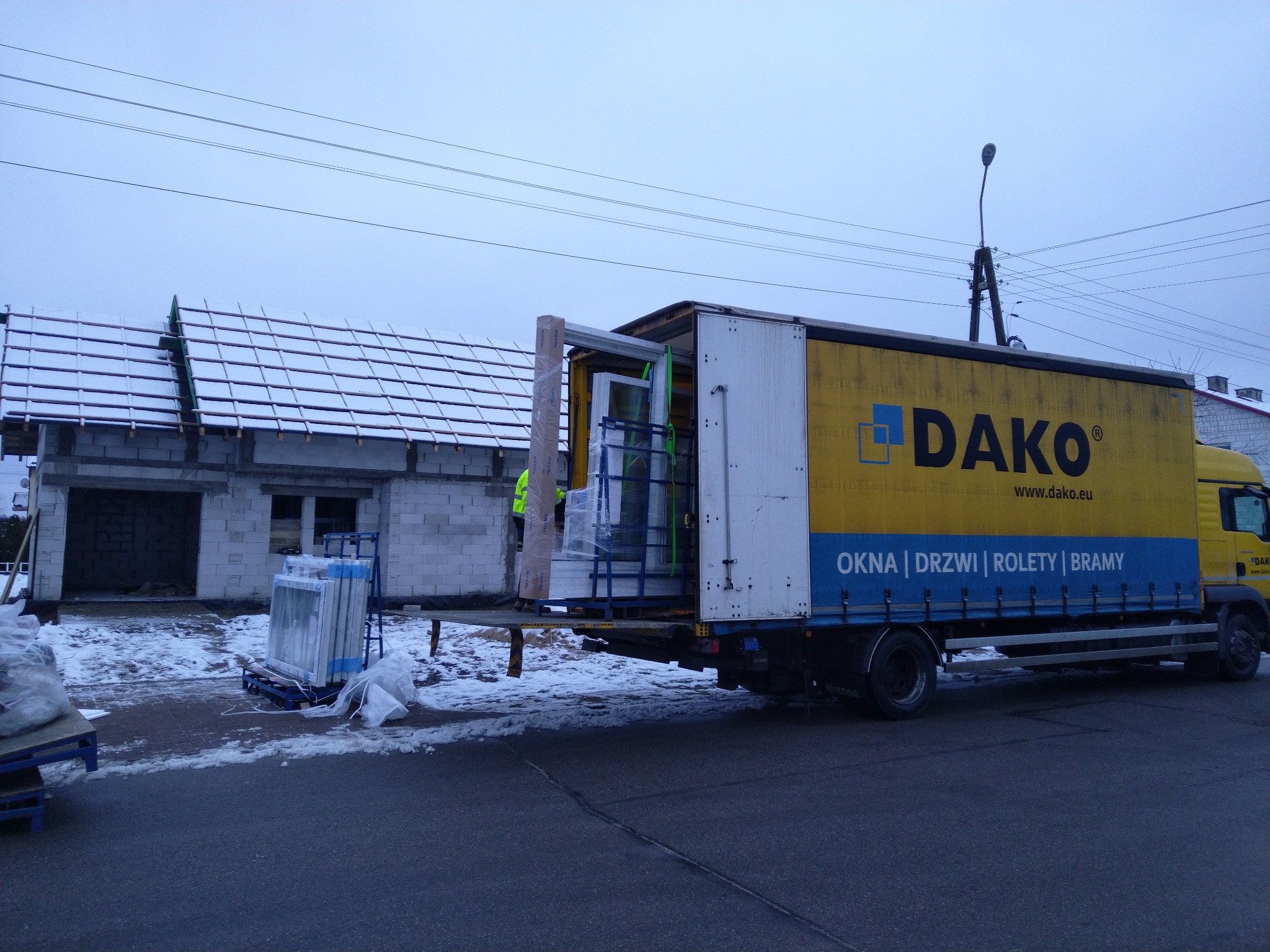 Żółto-biała ciężarówka firmy Dako z otwartą tylną klapą, załadowana oknami, stoi obok budynku w stanie surowym zimą.