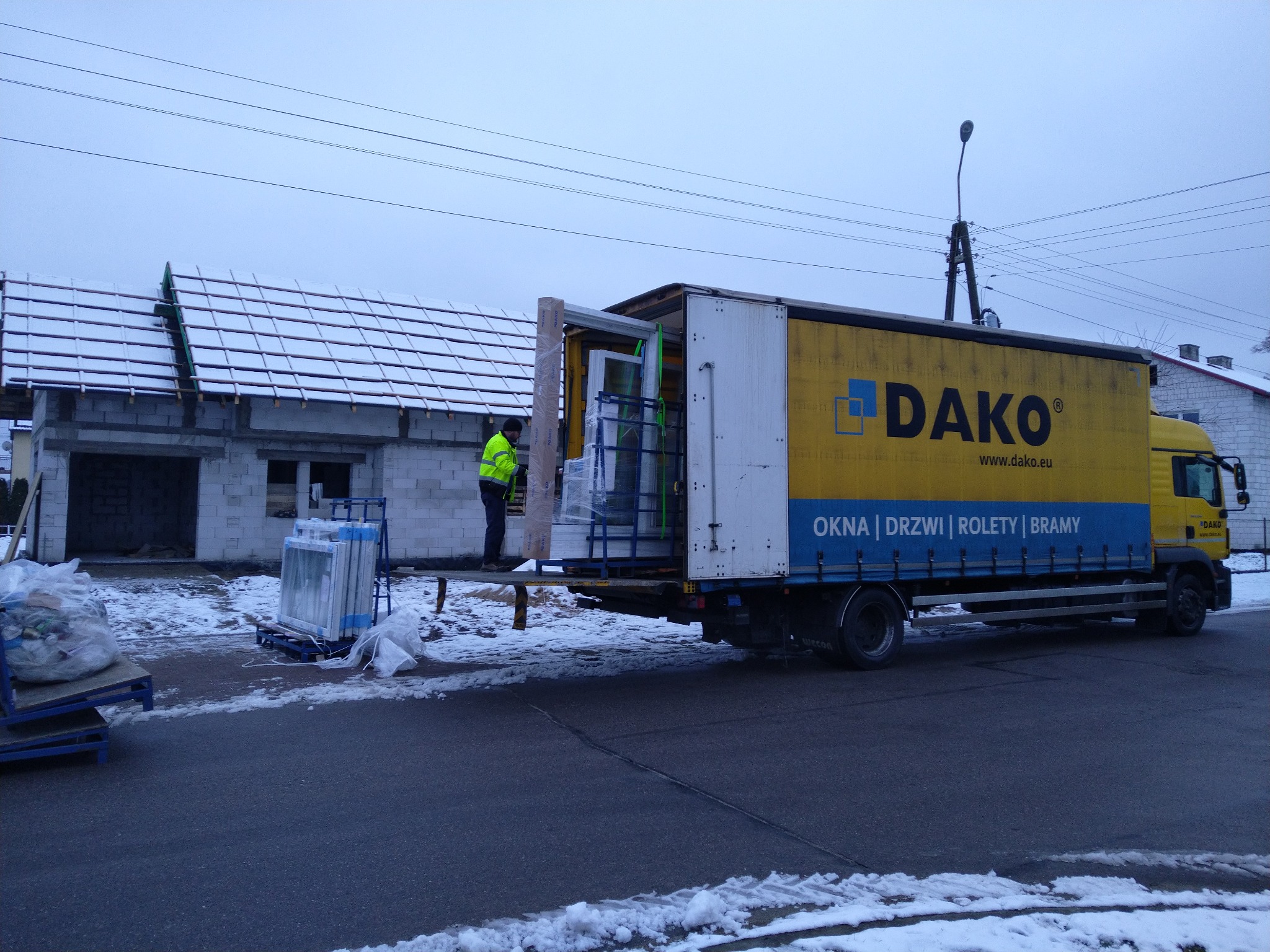 Dostawa okien na budowę: pracownik w żółtej kamizelce rozładowuje z ciężarówki okna zabezpieczone folią, obok stoją palety z kolejnymi oknami, w tle budynek w stanie surowym zimą.
