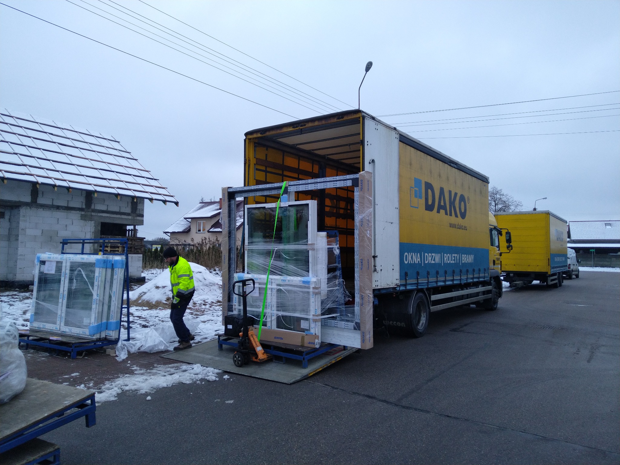 Rozładunek pakietów okiennych z samochodu dostawczego na budowie w zimowy dzień, pracownik w odblaskowej kamizelce, w tle budynek w stanie surowym i ośnieżone dachy.