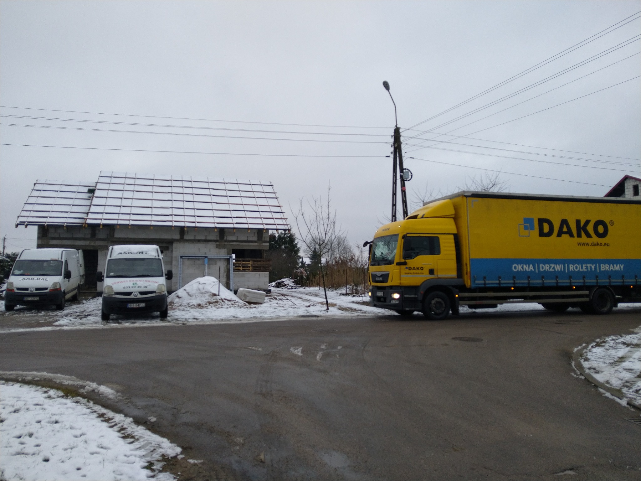 Żółta ciężarówka z napisem DAKO (okna, drzwi, rolety, bramy) na tle budowy domu w zimowej scenerii, z dwoma zaparkowanymi białymi busami i stertą śniegu.