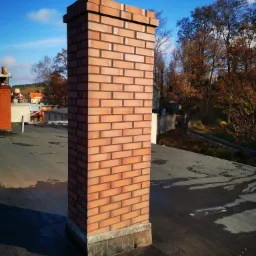 Nowy, murowany komin z cegły klinkierowej na czarnym, płaskim dachu z widocznymi śladami wody, w tle zabudowania i drzewa pod błękitnym niebem.