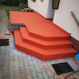 Pomarańczowe schody wejściowe do budynku, zabezpieczone niebieską taśmą malarską, z widocznymi śladami farby na kostce brukowej.