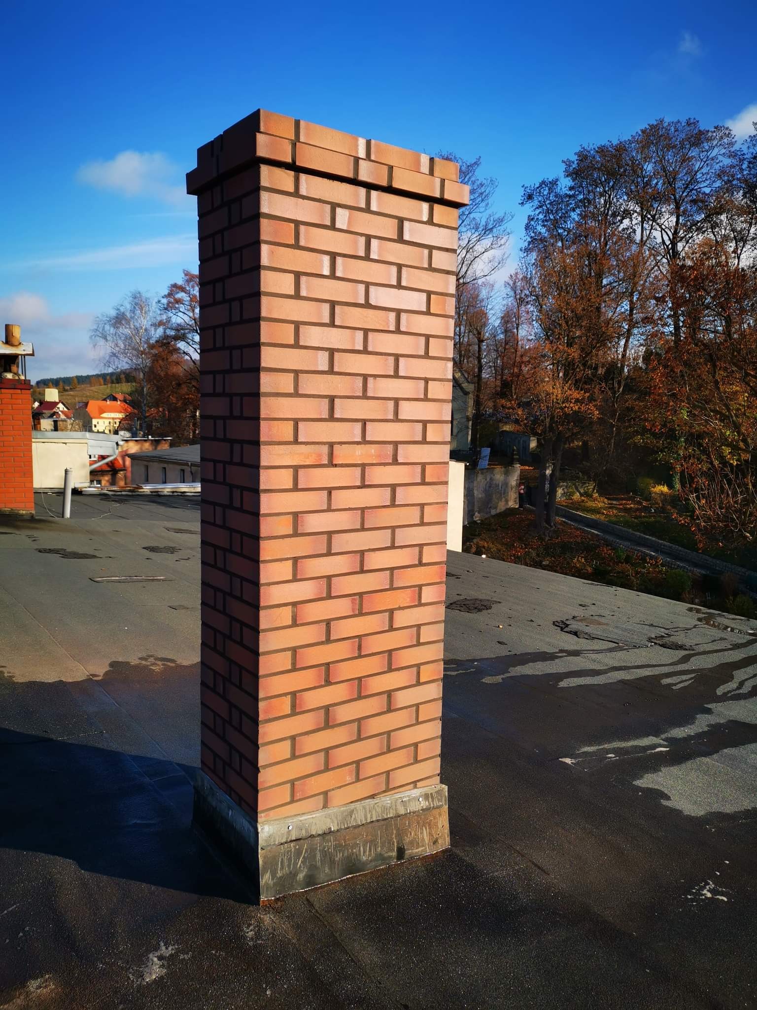 Nowy, murowany komin z cegły klinkierowej na czarnym, płaskim dachu z widocznymi śladami wody, w tle zabudowania i drzewa pod błękitnym niebem.