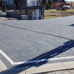 Artur Bąbiak - Ułożona ciemnoszara kostka brukowa z białymi liniami oddzielającymi, obrzeża z szarego betonu, z zamontowanym odpływem liniowym, na posesji z ogrodzeniem z czarnych sztachet i bramą.