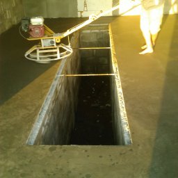 Artur Bąbiak - Praca w toku: Wygładzanie wylewki betonowej za pomocą zacieraczki mechanicznej, widoczny fragment stopy pracownika bez obuwia oraz otwór w podłodze zabezpieczony prętami.