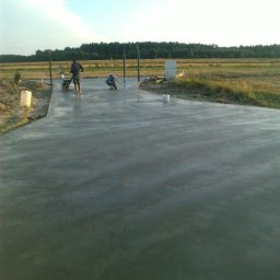 Artur Bąbiak - Świeżo wylana, gładka posadzka betonowa na zewnątrz, dwóch pracowników w trakcie wygładzania powierzchni, widok na pola i las w tle.