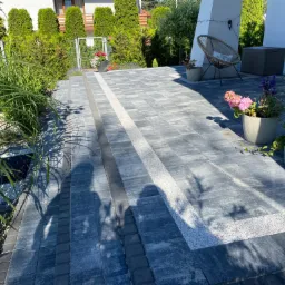 Nowoczesny taras z szarej kostki brukowej z jasnymi pasami dekoracyjnymi, widok na dom z tujami i wiklinowy fotel.