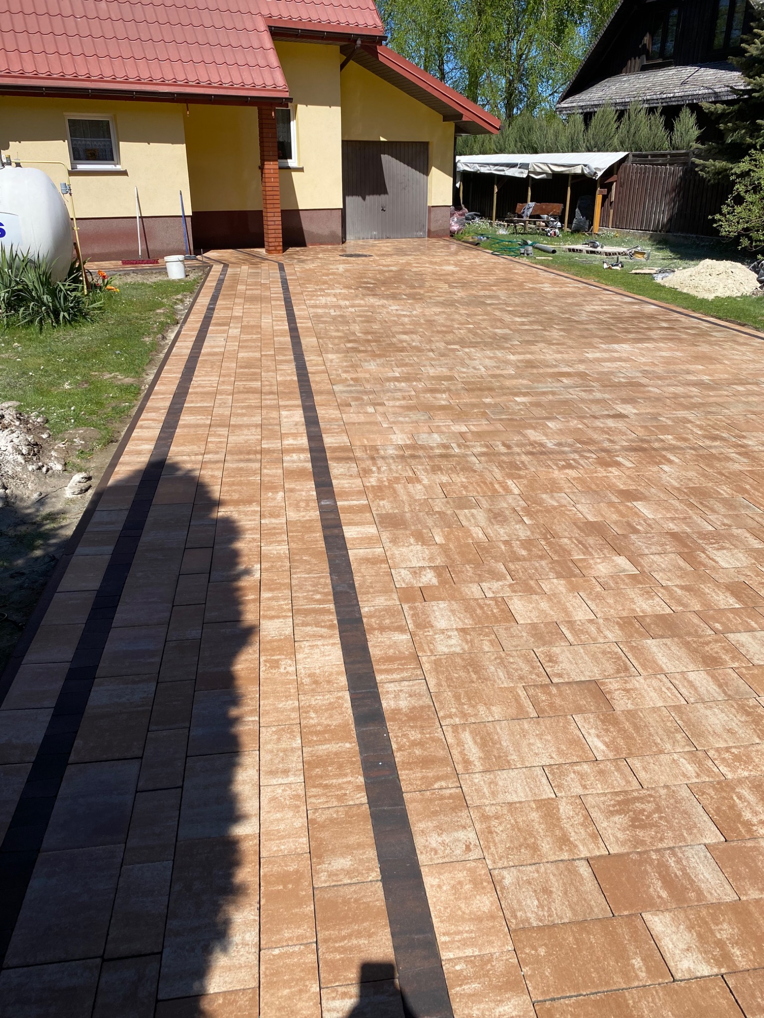 Nowo ułożony podjazd z kostki brukowej w odcieniach brązu i grafitu, prowadzący do domu z żółtą elewacją i czerwoną dachówką w słoneczny dzień.