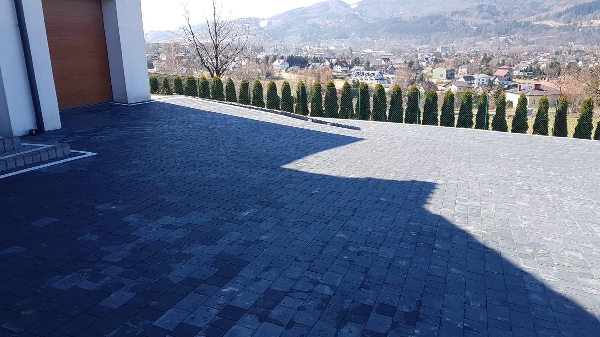 Nowoczesny podjazd z ciemnej kostki brukowej ułożonej we wzór, z widokiem na góry i rząd tuj za metalową siatką.