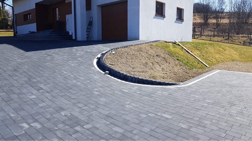 Nowo ułożona, szara kostka brukowa prowadząca do domu z garażem, obrzeża z ciemnych kostek oddzielają ścieżkę od trawnika z nasypem ziemi.