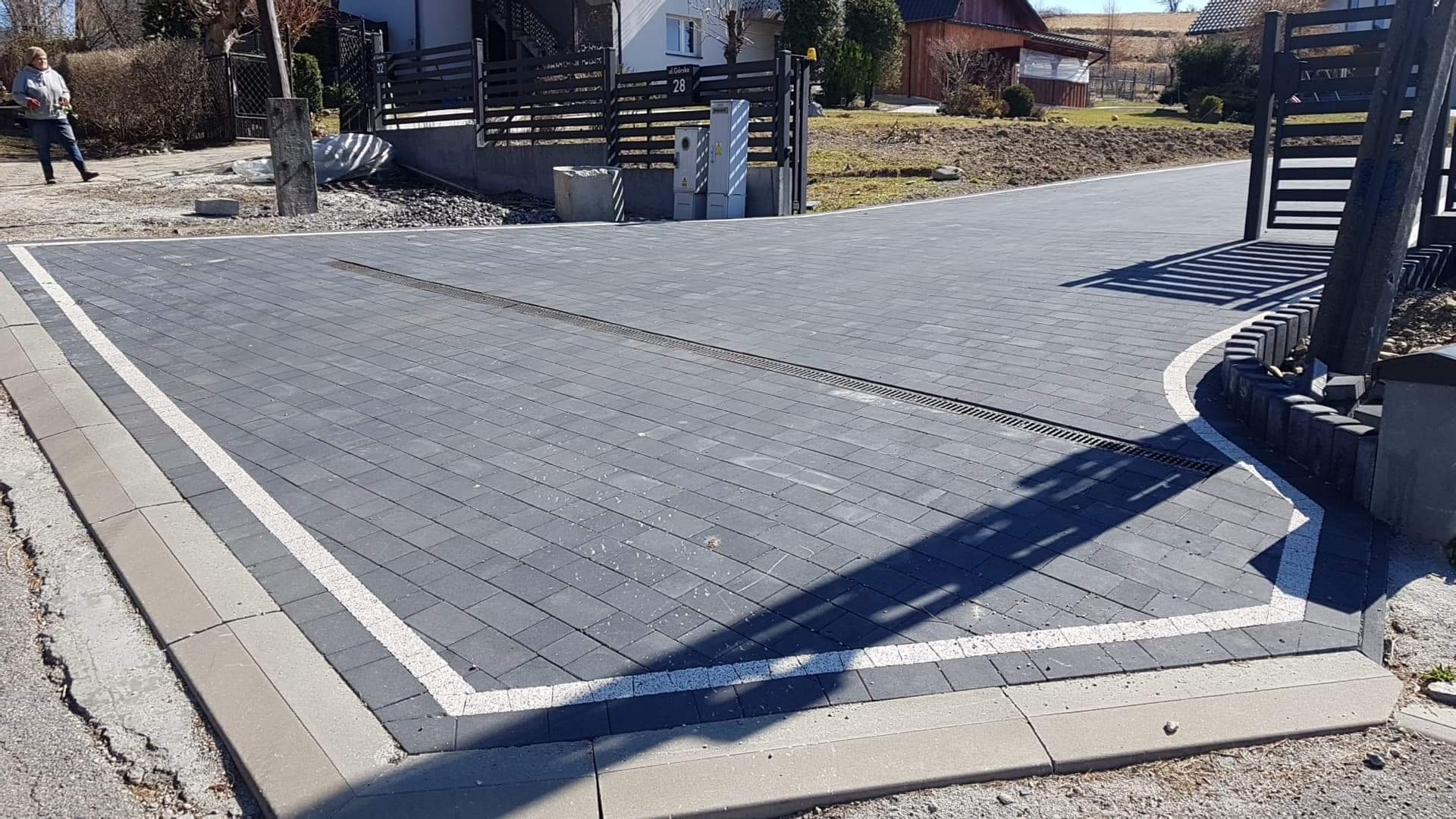 Ułożona ciemnoszara kostka brukowa z białymi liniami oddzielającymi, obrzeża z szarego betonu, z zamontowanym odpływem liniowym, na posesji z ogrodzeniem z czarnych sztachet i bramą.