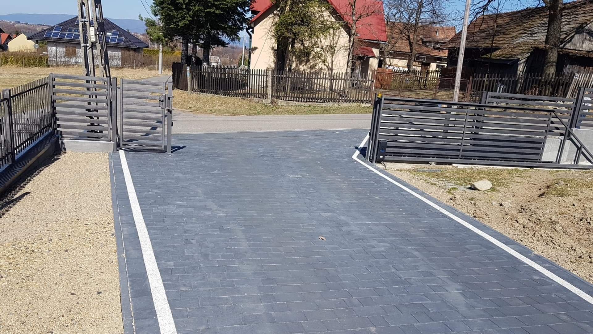 Nowo ułożona, ciemnoszara kostka brukowa podjazdu z białymi obrzeżami, prowadząca do nowoczesnej, metalowej bramy wjazdowej z poziomymi szczeblami, widok na okoliczne domy i drzewa.