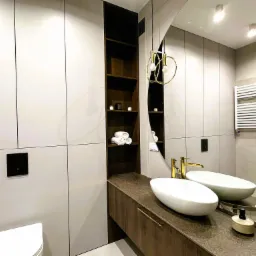 Nowoczesna łazienka z zabudową meblową w odcieniach szarości i brązu, owalna umywalka nablatowa, złota armatura, oświetlenie wiszące z elementami złota, otwarte półki z ręcznikami.