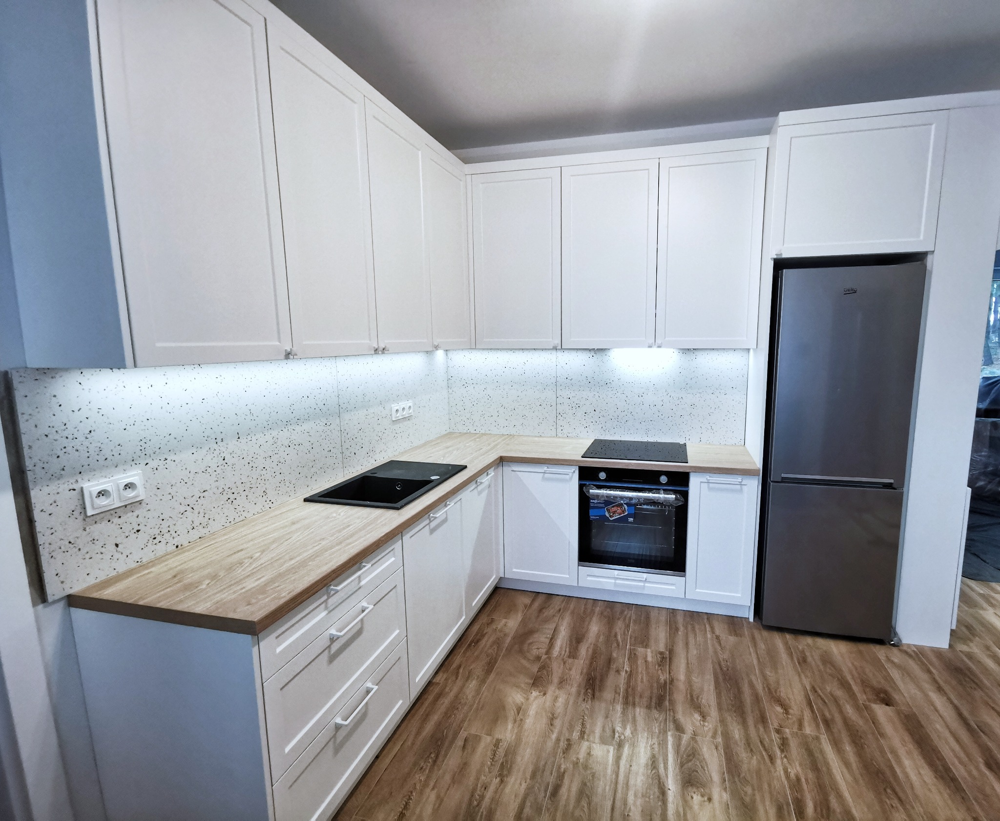 Nowoczesna kuchnia z białymi frontami, drewnianym blatem i szarym sprzętem AGD, oświetlenie LED pod szafkami wiszącymi, widoczny fragment podłogi imitującej drewno.