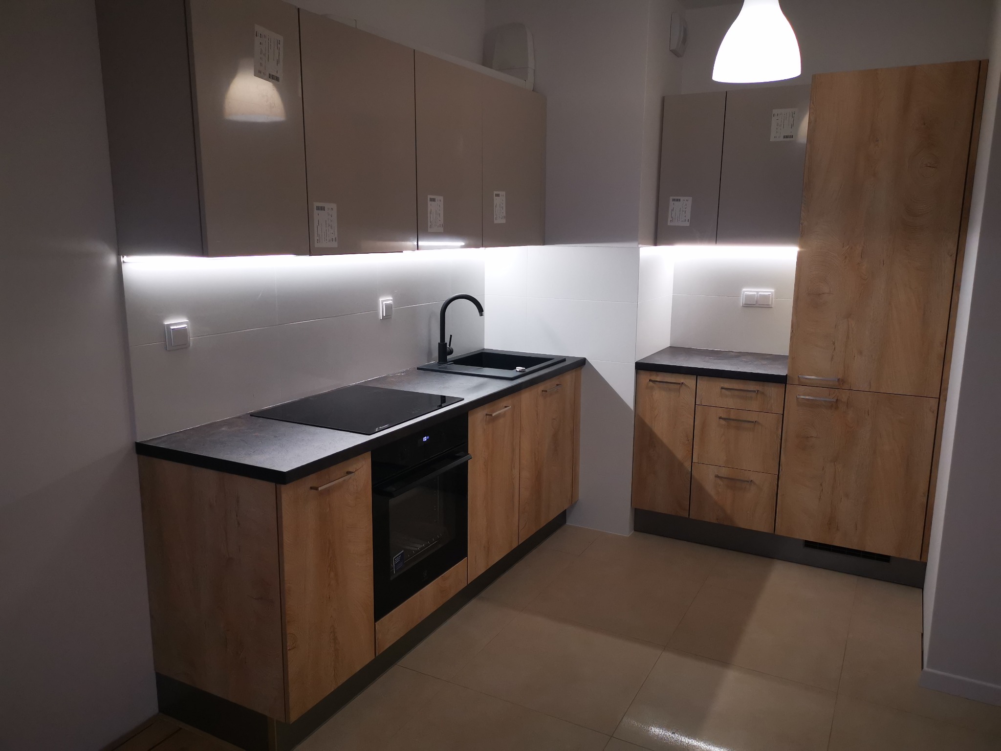 Nowoczesna kuchnia z drewnianymi frontami dolnych szafek i szarymi górnymi szafkami, czarny zlew i płyta indukcyjna, oświetlenie LED pod szafkami górnymi.